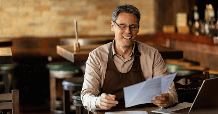 Man wearing apron sits at table in empty restaurant, smiling while holding papers.